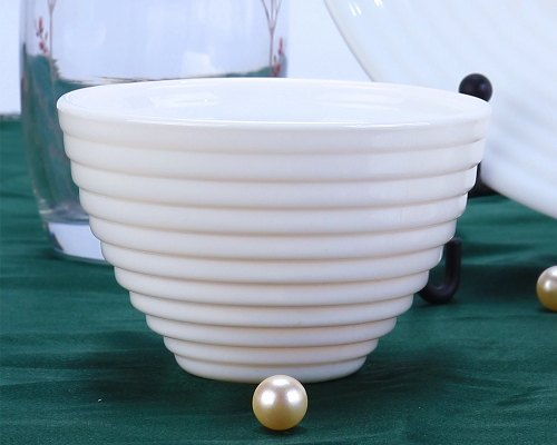 A creamy-white opal glass bowl with a ribbed, beehive design sits on a dark green surface. A single golden pearl is placed in front of the bowl.